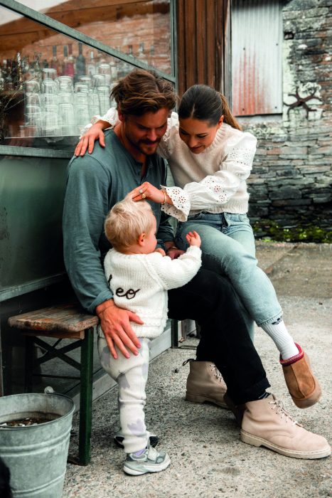 Picture of Louise, her fiance and her son Leo sitting outside on the bench.  They look happily at each other, with their arms around each other.  The background is of what appears to be a rustic barn, but is not fully visible.