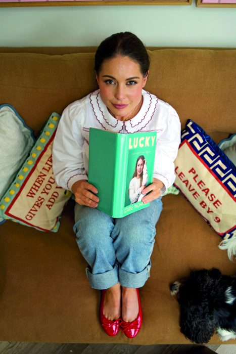 Louise is sitting on a brown couch holding her new book.  The book is green.  She wears a white blouse and blue jeans.  Her hair is pulled back in.  She smiles at the camera. 