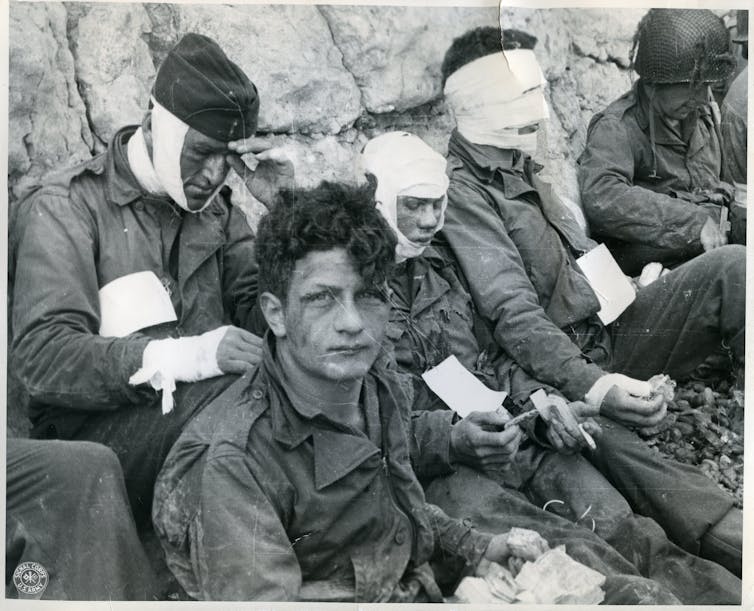 Men in uniform wearing bandages on their heads, chests and arms stand by a rock wall.